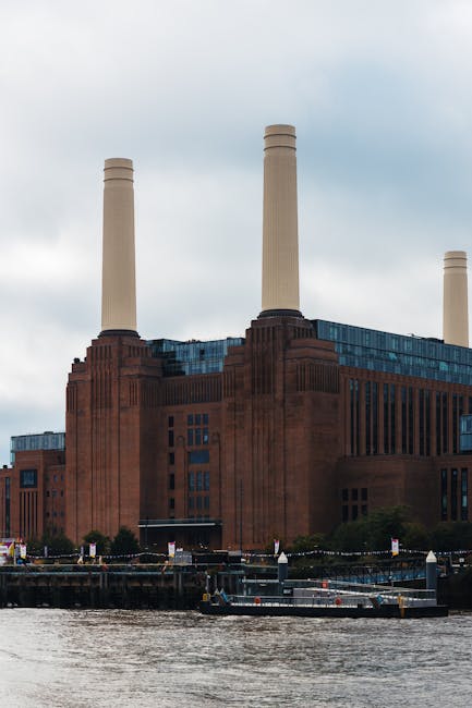 A large industrial building with a predominantly red brick facade, featuring vertical architectural details and rectangular windows in multiple rows. The structure spans horizontally across the image and is situated near a waterway, with a river or canal visible in the foreground. The building has several tall, cream-colored chimneys rising from its roof, some of which are grouped together, and the chimneys appear to be made of smooth concrete or similar material. The background shows a cloudy, overcast sky, while the environment around the building includes a dock or quay area with small boats and maritime equipment, indicating recent activity related to waste or material transportation, characteristic of industrial sites that may handle waste management or other bulk cargo. The image, as part of a waste management or rubbish removal context, could relate to the industrial handling of waste products or materials requiring external collection services, exemplifying alternative waste handling options outside typical residential rubbish collection.