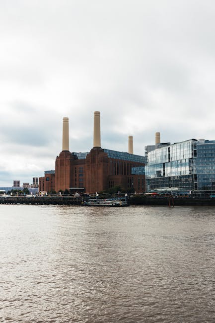 A large industrial building with three tall, beige smokestacks emerging from its dark red brick facade, situated along the riverbank. To the right of the historic structure, there is a modern, glass-fronted office building reflecting the cloudy sky, with multiple stories and a sleek, contemporary design. The buildings are positioned on the edge of the water, with a wide river in the foreground showing gentle ripples on its surface. The overcast sky has a mix of grey and white clouds, casting diffuse light over the scene. The overall environment suggests a riverside urban area, where historic and modern architecture coexist, typical of a city scene that may involve private waste handling or on-site clearance for commercial or industrial premises, aligning with themes of rubbish removal and alternative waste management solutions provided by companies like Waste Removal Battersea.