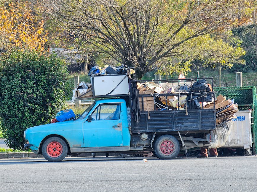 A small vintage blue pickup truck with rusted red wheels is parked along a street with a row of trees in the background. The truck's cargo bed is filled with an assortment of waste materials, including cardboard boxes, black plastic bags, and miscellaneous debris, which are loosely stacked and secured with ropes. The truck is positioned next to a green fenced area and appears to be used for private rubbish collection or on-site waste removal, as it is heavily loaded with bulky waste. The surrounding environment includes a sidewalk, a dense green bush on the left, and a tall tree with both yellow and green leaves behind the vehicle, suggesting an autumn setting. Natural daylight illuminates the scene, highlighting the textures of the materials and the weathered surface of the truck, reinforcing its role in independent waste handling and rubbish clearance services in the area.