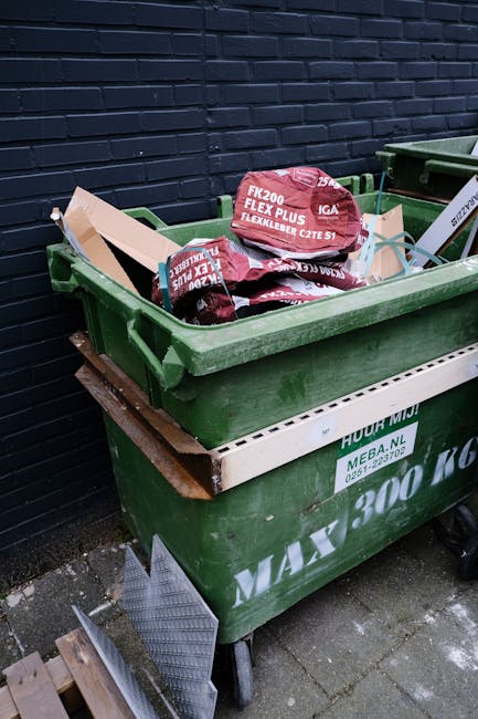 A large green wheeled waste container filled with red and black bags of garden or building waste, placed on a paved surface adjacent to a dark brick wall. The bags appear to contain soil, mulch, or similar materials, and are branded with white and red text. The container is positioned next to another similar container and on top of a wooden pallet, suggesting it is used for private or independent waste collection services. Nearby, there are small metal grid pieces leaning against the side of the container, and the area appears to be an outdoor storage or disposal zone, consistent with waste removal activities typical of private rubbish services such as those offered by Waste Removal Battersea.