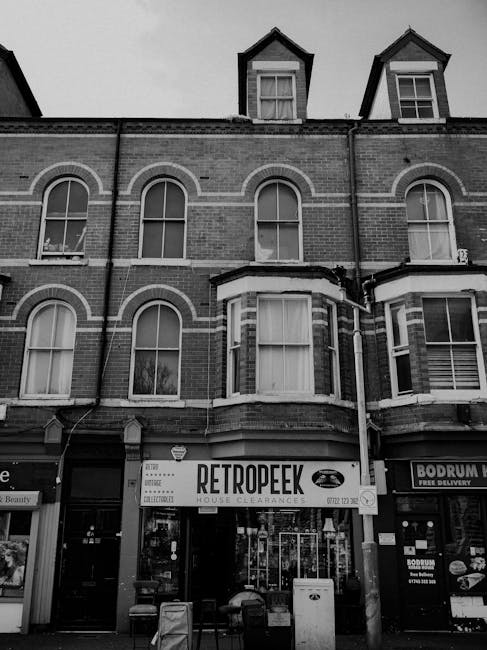 The image shows the façade of a multi-storey brick building with Victorian-style architecture, featuring large arched windows on the upper floors and several dormer windows on the roof. The ground floor houses a retail unit with a prominent sign reading 'RETROPEEK' advertising house clearances. In front of the shop, there are two benches, a small waste receptacle, and a mailbox, all positioned on a paved pavement. Adjacent to the retail unit, other commercial signs are visible, including one for a delivery service. Visible external drainpipes and wiring run along the building's façade. The scene appears to be captured during daylight hours with overcast weather, contributing to the monochromatic, grayscale tone of the image. This setting, by illustrating a typical urban commercial frontage, subtly relates to private waste handling and alternative waste collection methods, such as those provided by waste removal services like Waste Removal Battersea, which handle clearance of household and commercial rubbish outside local authority provision.