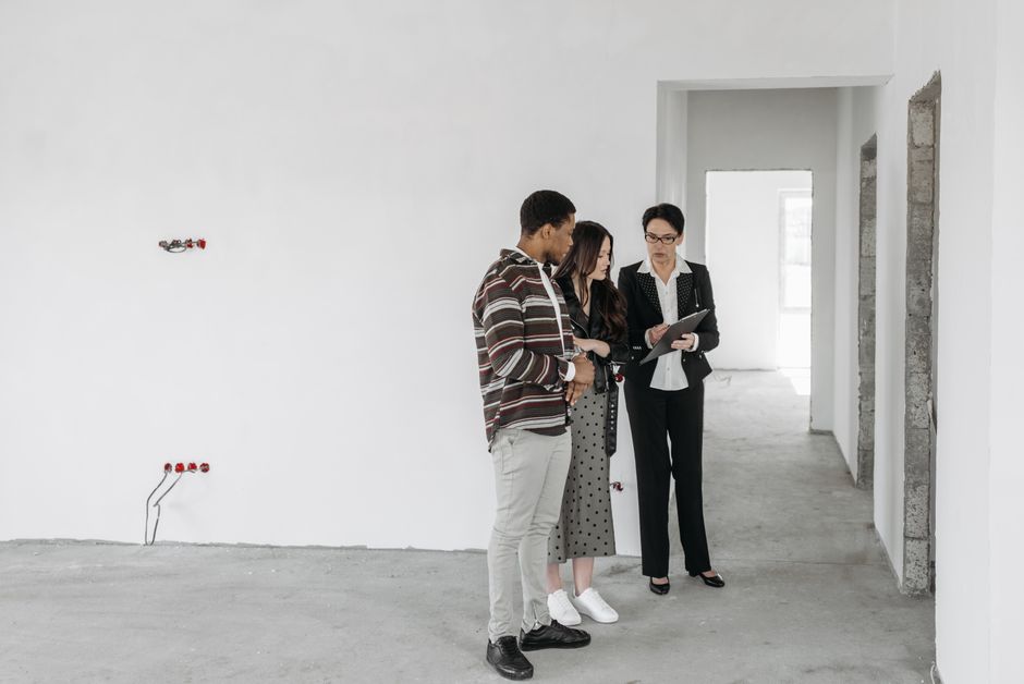 Three individuals, two women and one man, are standing indoors in an unfinished room with exposed concrete and plaster walls. The woman on the right, dressed in a black business suit with a white shirt, is holding a tablet and appears to be explaining something to the other two, who are listening attentively. The man on the left is wearing a striped brown and white jacket with light grey trousers, while the woman in the middle has long dark hair, wears a black top with patterned grey trousers, and white sneakers. The room is well-lit with natural light coming through a window at the far end, and the walls are mostly white but have visible brickwork and construction gaps in some areas indicating ongoing renovation or building work. The scene may relate to building or property inspection, potentially linked to private estate clearance or refurbishment, with a context of professional oversight for upcoming rubbish removal or interior renovation—services often handled by companies like Waste Removal Battersea.