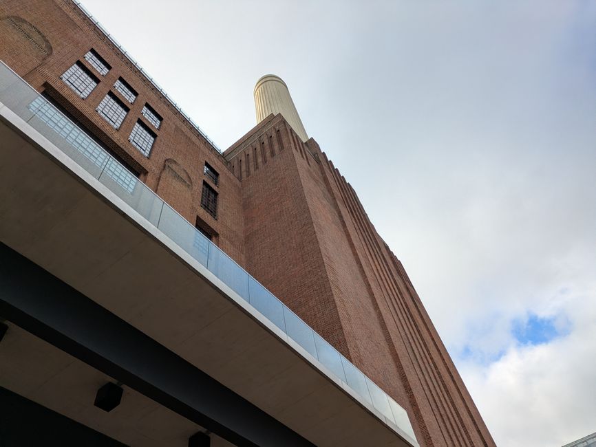 A low-angle view of a historic brick power station building with a tall, cylindrical chimney extending upwards into a cloudy sky. The building’s exterior features red-brown brickwork with evenly spaced rectangular windows, some with black metal grilles. A modern glass and metal railing lines a small balcony or walkway on the building's lower section. The scene is outdoors, likely in an urban environment, with natural daylight illuminating the textured brick surface and the contrasting smooth glass elements. The perspective emphasizes the scale and industrial character of the structure, which is relevant to site clearance or alternative waste handling considerations by Waste Removal Battersea providing independent rubbish removal services near Battersea Power Station.