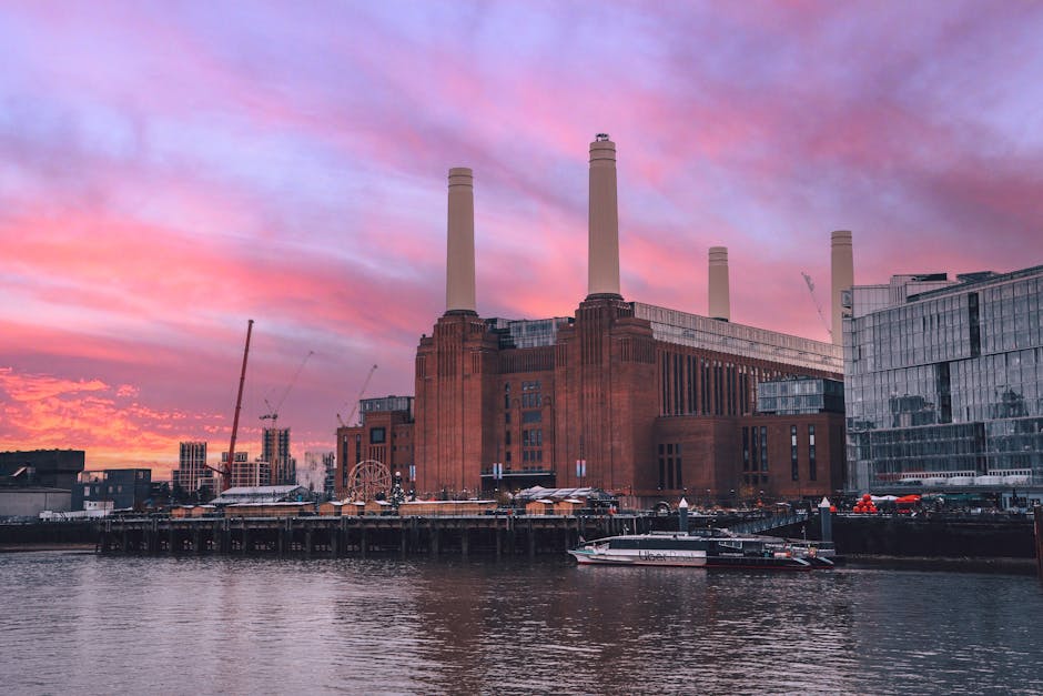 A view of the Battersea Power Station with its iconic four tall chimneys during sunset. The sky is painted with pink, purple, and orange hues, casting a warm glow over the red brick building. In the foreground, the River Thames reflects the colorful sky and the structure. Along the riverbank, there is a dock area with small boats and a pier. To the right of the power station, modern glass buildings are visible, contrasting with the historic industrial architecture. Construction cranes are seen in the background, indicating ongoing development. The scene showcases a blend of historical industrial architecture and contemporary urban growth, with the power station serving as a prominent landmark in the Battersea area. The lighting highlights the textures of the brickwork and the reflections on the water, evoking a tranquil yet active riverside environment relevant to the context of property and site management, often associated with urban waste and rubbish management preparations near such iconic landmarks. Waste Removal Battersea might handle rubbish clearance in this vibrant riverside area, supporting private waste management solutions rather than council-led disposal.