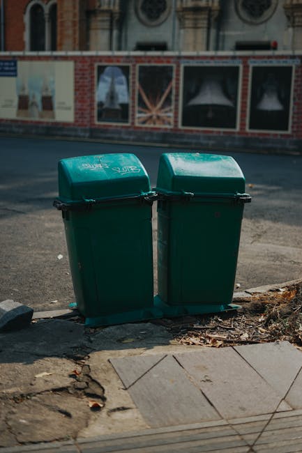 Two green plastic rubbish bins with hinged lids are positioned on a concrete pavement at the edge of a street, with the left bin slightly leaning inward. The bins are made of durable, textured plastic and are marked with white lettering, indicating their use for waste collection. They sit on a small black plastic tray to catch any runoff, and the surrounding pavement shows cracks and scattered dried leaves. Behind the bins, there is a curb separating the pavement from the asphalt road, which features visible tire marks and appears well-used. In the background, an urban environment is visible with a brick building that has decorative window frames and large display windows covered by protective or promotional posters, slightly reflective in the daylight. The overall scene suggests an area where private waste disposal or alternative rubbish collection might be employed, overseen by waste removal services such as Waste Removal Battersea, to support local rubbish management needs.